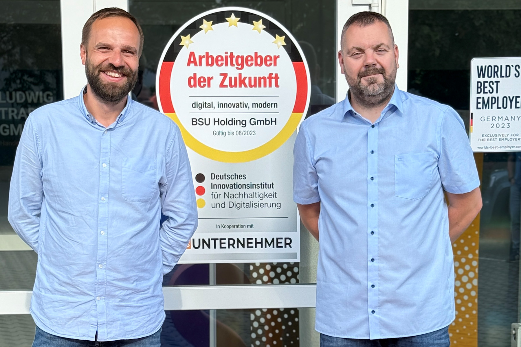 Die neuen Geschäftsführer der BSU Holding, Matthias Gommlich und André Tischler, stehen vor dem Firmensitz mit großem BSU-Logo und Auszeichnung ‚Arbeitgeber der Zukunft‘.
